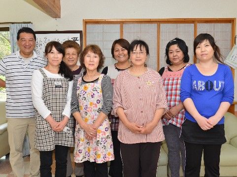 施設の壁をバックに8人のスタッフが笑顔で立っているのが写っている。男性1名と女性7名。年齢層は幅広いスタッフが在籍していることが判る。