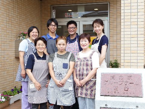 ベージュ色のタイル調の外壁の玄関前。施設名の看板の横に前列に3人と後列に4人のスタッフが並んでいる。