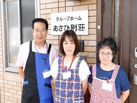 玄関出入口の施設名の表札前でスタッフが3人並んでいる。3人ともそれぞれ違った柄のエプロンを掛け、名前札を掛けている。