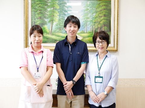 白壁に掛けられた大きな絵画の前にスタッフ3人が並んでいる。真中に男性がいて両側に女性がいて、皆笑顔である。