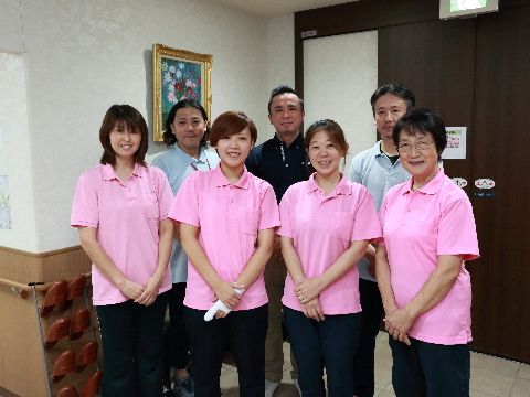 施設のスタッフ達が笑顔で2列になって並んでいる。女性4人、男性3人の合計7人がいる。女性は同じ服を着ている。