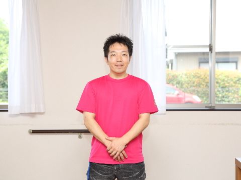 男性の職員が一名、窓の前に立って写真に写っている。窓にはレースのカーテンが取り付けてある。紅い車が一台停まっているのが見える。