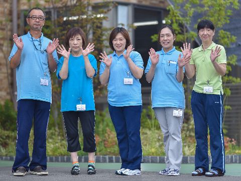施設で働く職員五人が写真に納まっている。左端に男性の職員が一人、残り四名は女性である。ユニフォームを着衣し、同じポーズで外に立っている。
