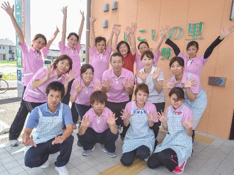 外壁に掲げられた施設名の前に15人のスタッフが3列に並んでいる。前2列は胸のあたりに両手を広げ、後部1列は手を挙げている。