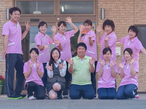 前方に5人、後方には6人のスタッフが並んでいる。グリーン色のユニフォームの男性と事務服の女性の他はピンク色のユニフォームを着用。