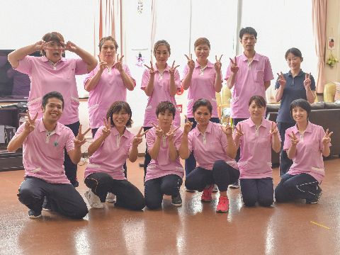 前列に6人、後列に6人のスタッフが並んでいる。ピンク色のユニフォームが印象的で、後列の端の女性だけ紺色のユニフォーム。