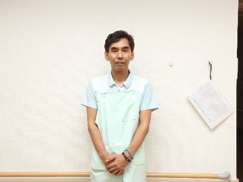 私服の白いポロシャツの上に、水色のエプロンを付けた男性スタッフの写真。施設内の手すりの付いた壁の前で撮影している。