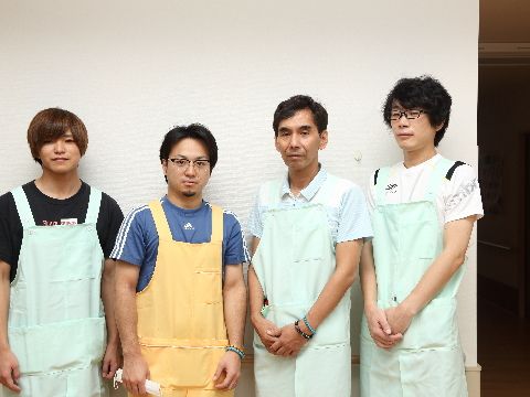 年齢の違う男性スタッフが施設内の真っ白な壁の前で、横一列の並んで写った写真。エプロンを着用している。