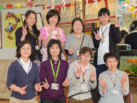 施設で働いている女性スタッフ8人が、前後2列に並んでいるエプロンやポロシャツなど、動きやすい格好をしている。