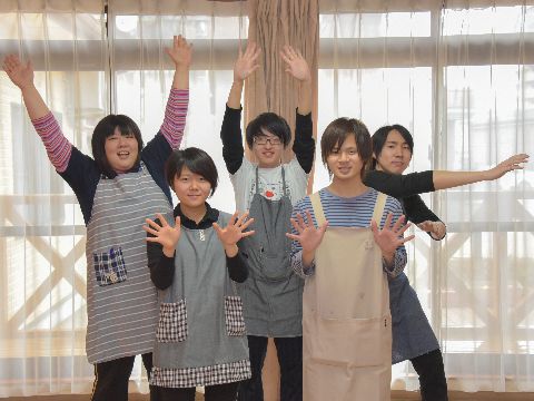 様々な年代の男女のスタッフが大きな窓をバックにしてポーズをとっている。スタッフは全員エプロンを着用している。
