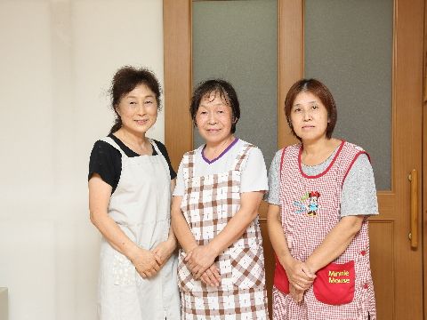 施設スタッフの紹介写真である。扉を背景にナチュラルメイクの女性が3人立っている。私服にエプロンを着用している。