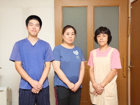 施設のスタッフを紹介する写真である。扉の前に1人の男性と2人の女性が立っている。半袖のTシャツを着用している。