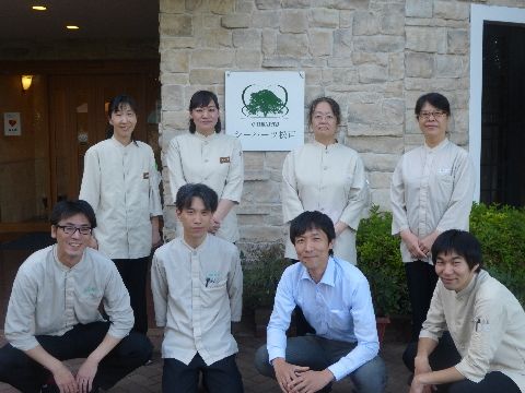 何人ものスタッフが施設の外で看板の前で写真に写っている。薄いクリーム色の制服を着たスタッフとブルーのシャツのスタッフが1人いる。