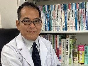 院長の写真。白衣姿でこちらを向いて座っている。後ろの本棚にはたくさんの医学書が並べられている