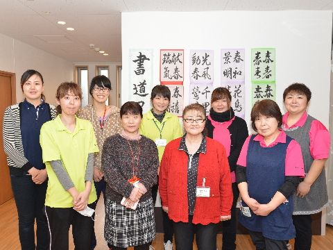 二列に並んで立つ笑顔のスタッフ一同。若者から年配者まで様々な年齢のスタッフがいる。壁には書道の作品が飾られている。