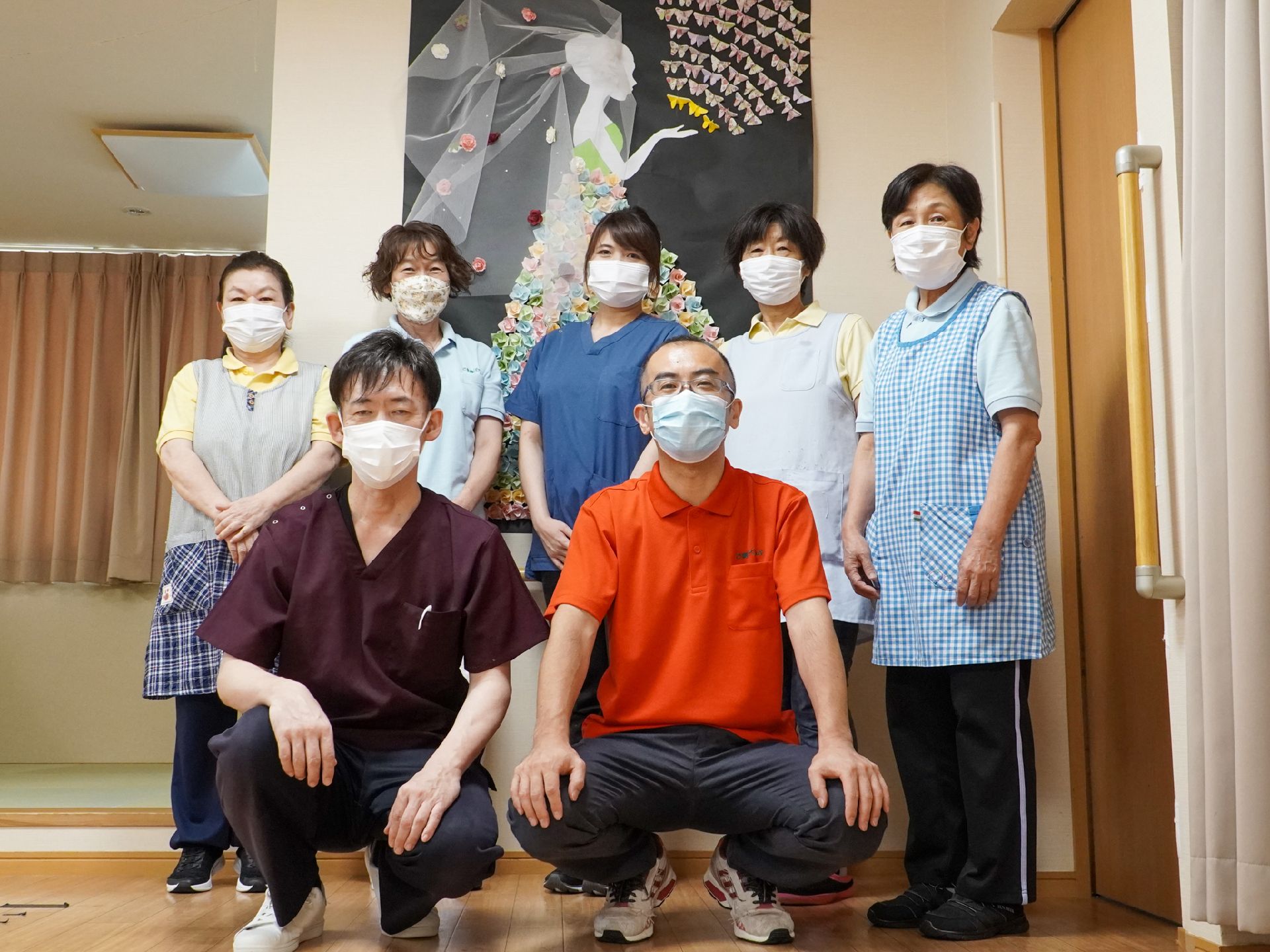 白い壁の前に立つ５名の女性従業員とその前にかがんでいる２名の男性従業員