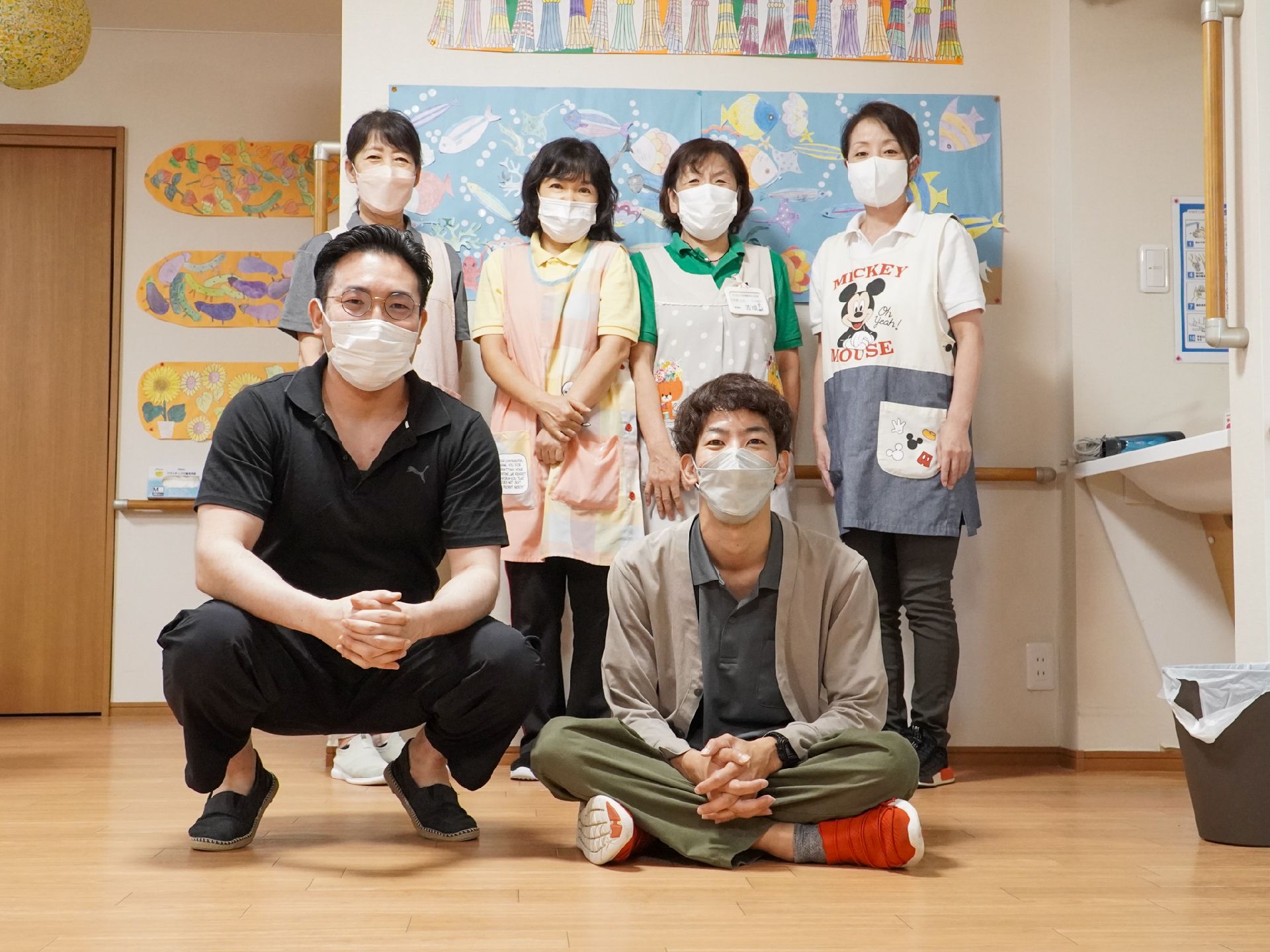 白い壁の前に立っている４名の女性従業員とその前に座っている２名の男性従業員