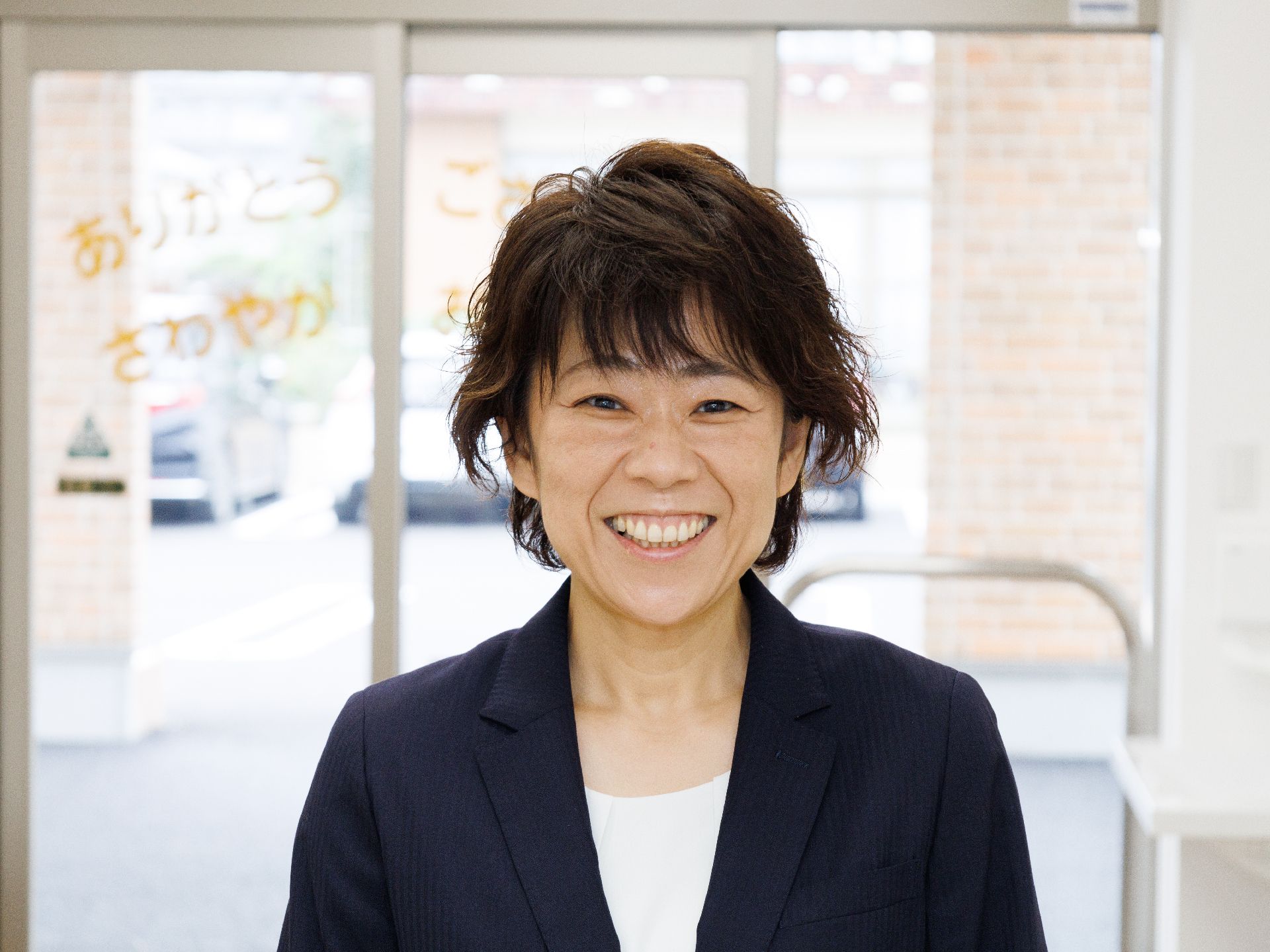 建物の入り口に配置された透明な扉の前に笑顔で立っているスーツを着た女性従業員