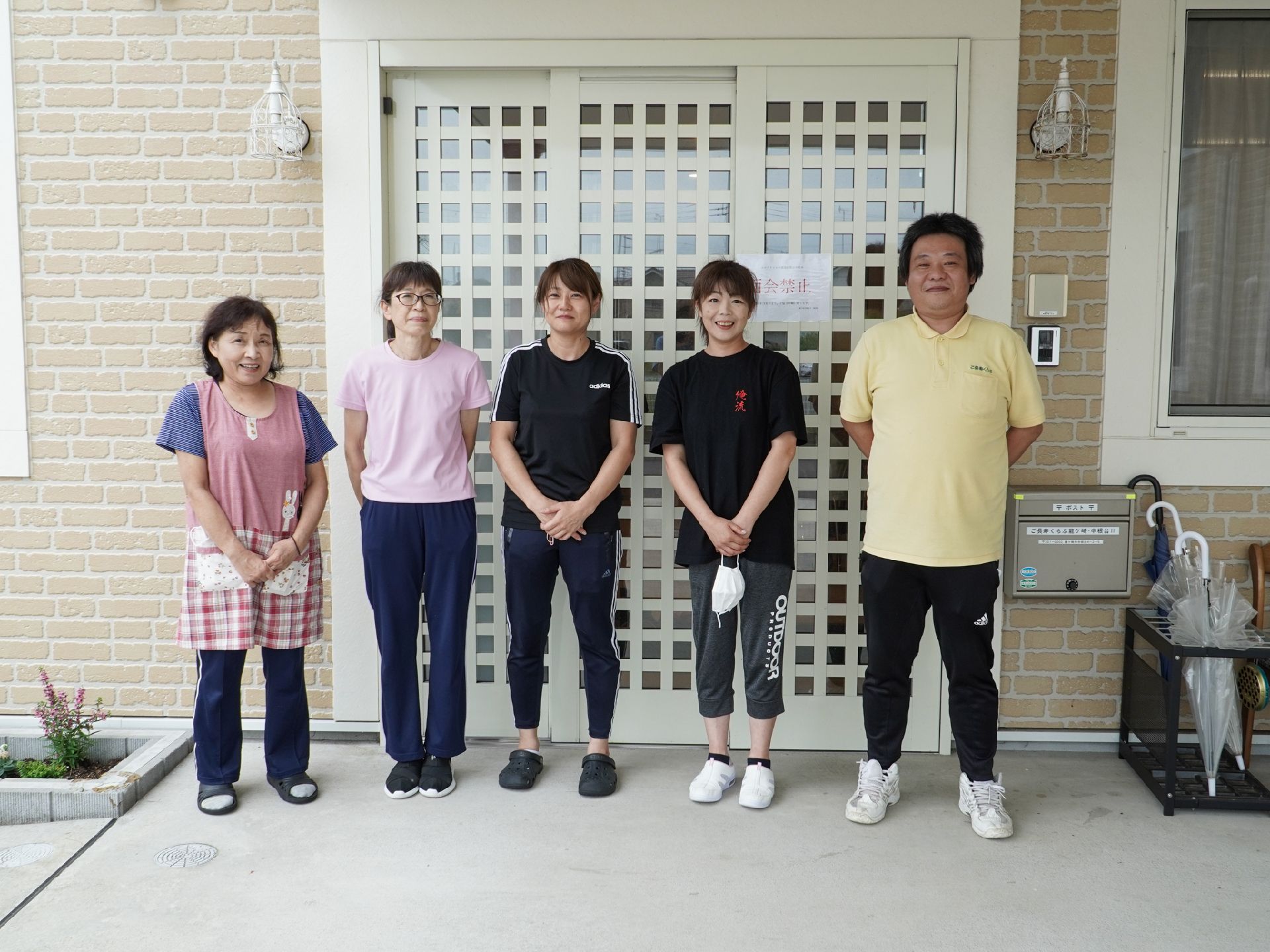 白い壁の建物の入り口前に並んで立っている５名の従業員