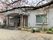 桜の花がきれいに咲いている枝が手前にあり、その奥に白い外壁の平屋建ての建物があり、玄関周りには花が植えられたプランターがたくさん置かれている。