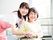 車いすの高齢女性とエプロンを付けた若い女性スタッフが寄り添い、笑顔で過ごしている。