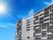 青空の下に建っている白を基調とした建物は９階建てで各フロアにベランダがあり、太陽の光が降り注いでいる。
