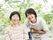 車いすの高齢女性に寄り添い、笑顔で過ごしているベージュのエプロンを付けた若い女性スタッフ。屋外の緑の多い場所で。