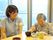 眼鏡をかけた女性入居者が食事をしているそばで、にこやかに見守る女性スタッフ。