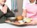 トレーに乗った食事を運んでいるエプロンを付けた若い女性スタッフと手を合わせて座っている高齢男性。