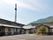 後方には緑あふれる山があり、前方には広い駐車場がある平屋建ての建物。車も止まっている。