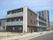 3階建ての建物はブラウン系でベランダ付き。周辺には高い建物や道路もあり、建物周辺にはのぼりが立っている。