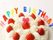 カラフルなアルファベットのロウソクで「HAPPY　BIRTHADAY」とつづられたイチゴのショートケーキ。ケーキ中央のイチゴには小さなピンクのリボンが乗っている。