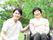 緑あふれる屋外で外気浴を楽しんでいる車いすの高齢女性と付き添いの若い男性スタッフ。