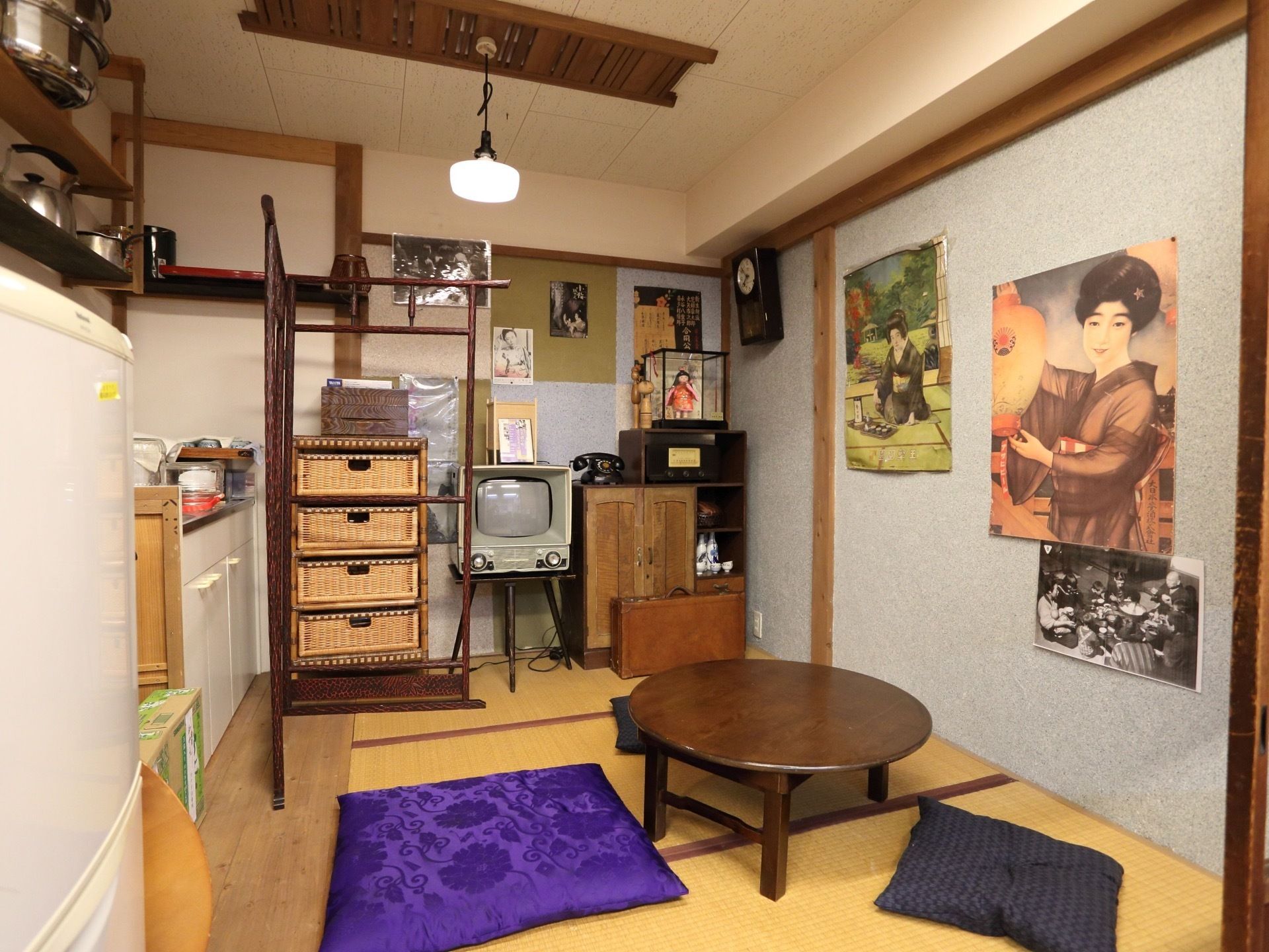 畳の上に丸いちゃぶ台があり、その周りに座布団が置いてある。右側の壁には、昭和レトロな雰囲気が漂うポスターが貼ってある。