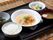 黒いお盆の上に、ごはん、みそ汁、高野豆腐の煮物、ニンジンやモヤシの入ったスープ料理、青菜の漬け物などが並んでいる。