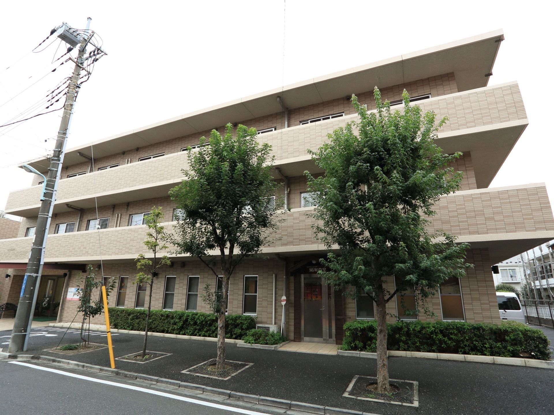 玄関の前方から、建物全体を写したものである。道路沿いには大きな木が植えてあり、左端には電柱が設置されている。