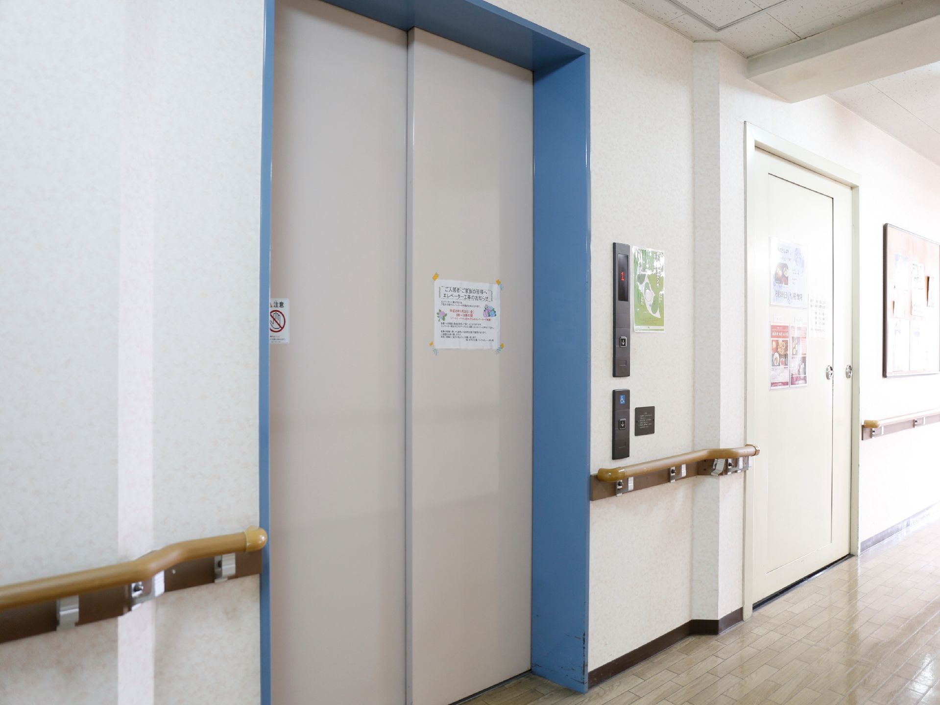 白いドアのエレベーター。三方枠は青く塗られている。ボタンが健常者用と身障者用に分かれている。奥に防火扉が見える。