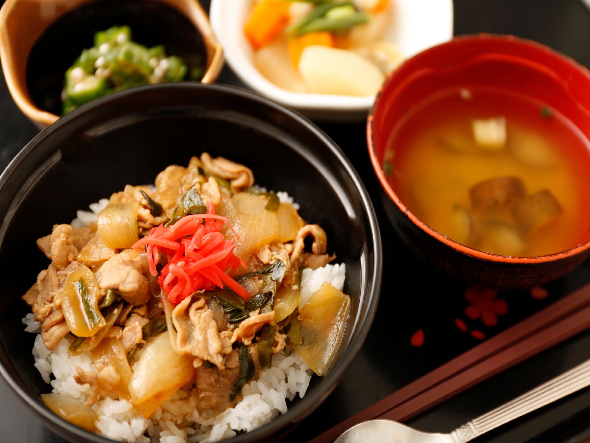 写真手前に大きく丼が映っている。玉ねぎ、肉などを煮た具の上に鮮やかな紅しょうがが乗った丼。周りに汁や小鉢、箸などが見える。