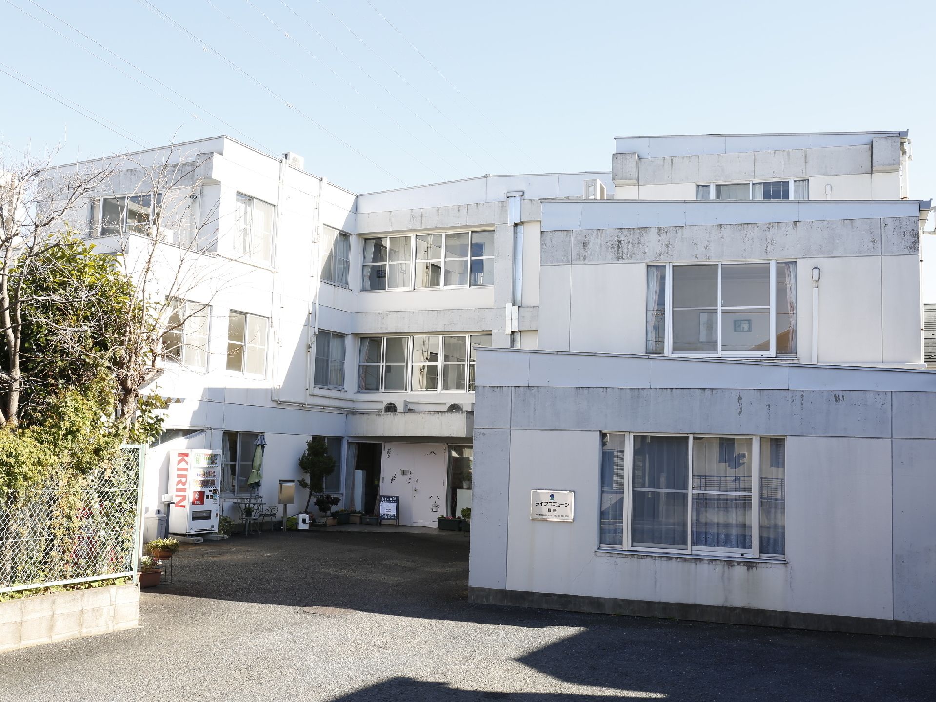 3階建ての白い建物。上に向かって階段状になっている。手前の壁に施設看板が設置されている。中央が凹んでおり、そこに玄関がある。