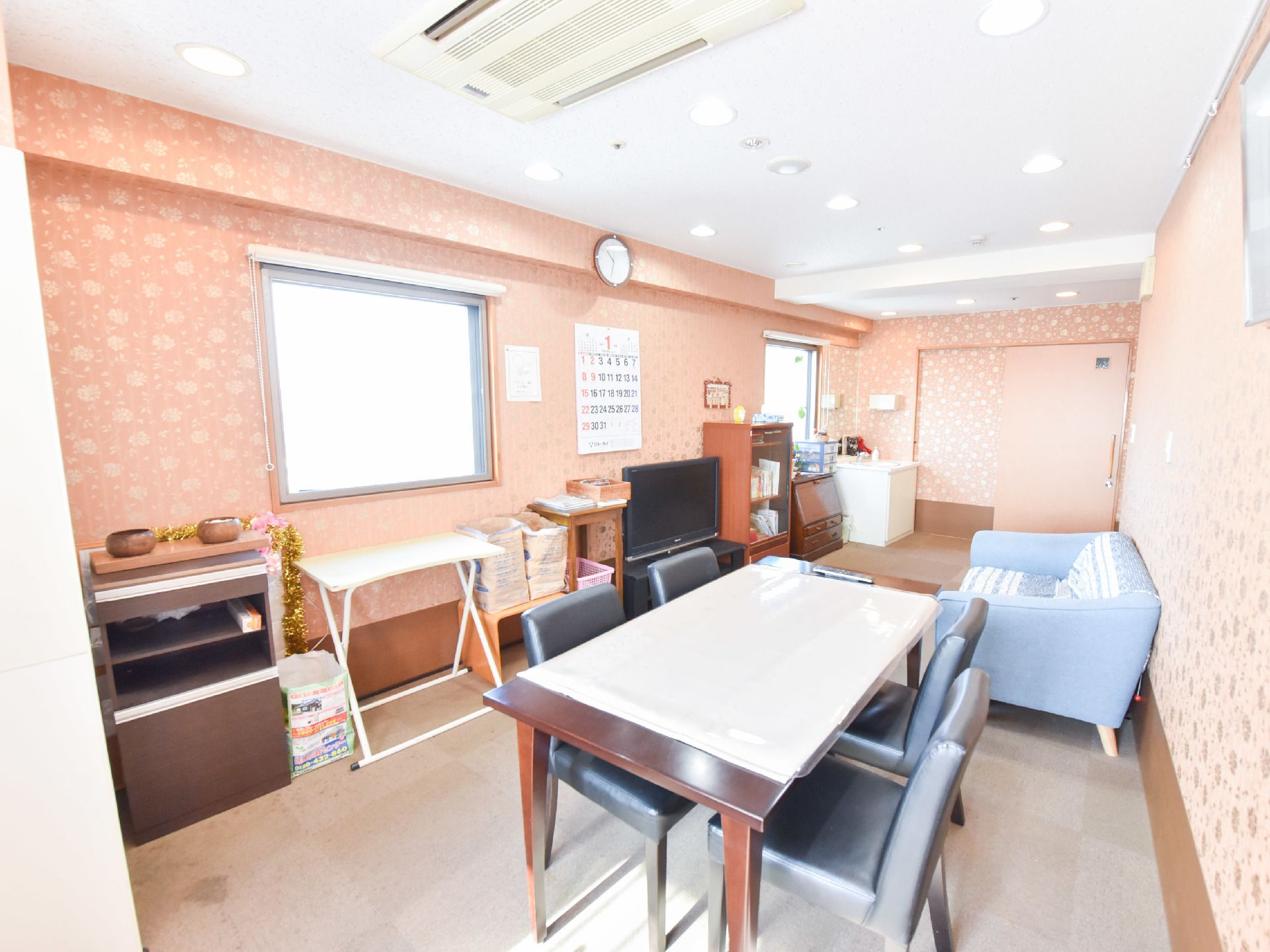 手前にはテーブルがあり椅子が4つ置かれている。壁には時計やカレンダーがあり天井には備え付けのエアコンが見える。壁際には棚が置かれている。