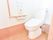 トイレは白壁に下部が茶色、床は木目のフローリング。水洗式トイレで壁には手すりとペーパー、トイレのスイッチが設置されている。