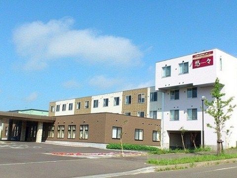 道路に近い部分が白色の外壁の３階建てになっている施設外観