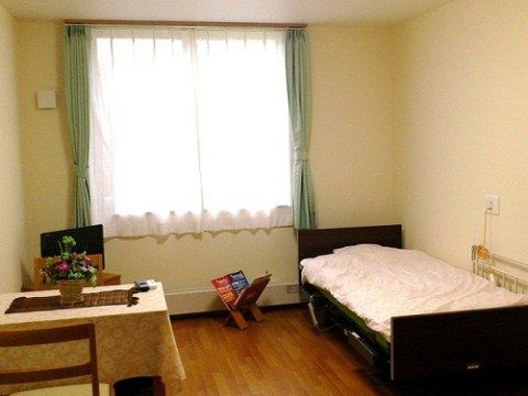 白を基調とした清潔感のある居室である。介護用ベッドや２人掛けのテーブルセット、ナースコール、カーテンが完備されている。