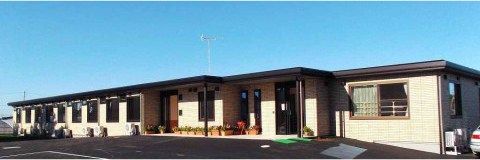 広い面積を有した、１階建ての落ち着いた色合いのグループホームである。施設前には駐車スペースを設けている。