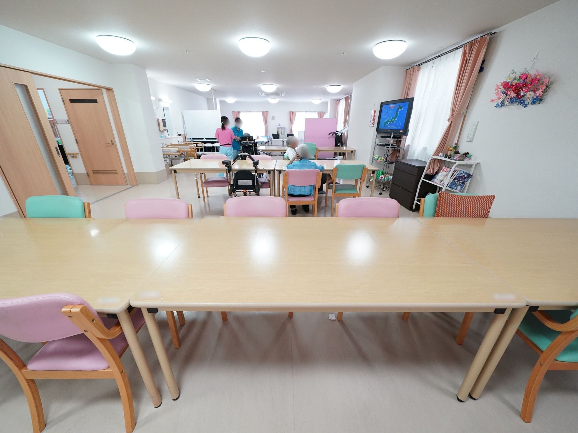 食堂には木製のテーブル、ピンク色や黄緑色の椅子が複数並べられており、席には入居者様が座っている。テレビも置かれている。