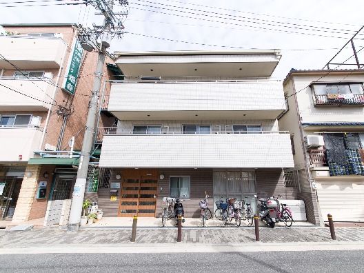 各フロアにベランダが付いた3階建ての建物。道路に面していて、両サイドに他の建物がある。