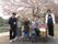 外で撮影された写真。桜の木の下に、５人の入居者様と２人のスタッフが並んでいる。向こう側には池も見える。
