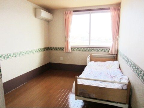 奥に窓があり、ピンク色のカーテンがついている。左側にエアコンが設置されていて、右側にベッドが置かれている。
