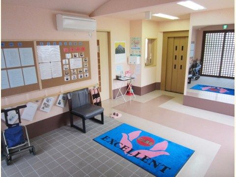 玄関はバリアフリー設計になっていて、鮮やかな色の玄関マットが敷かれている。左側に椅子やスリッパが置かれている。