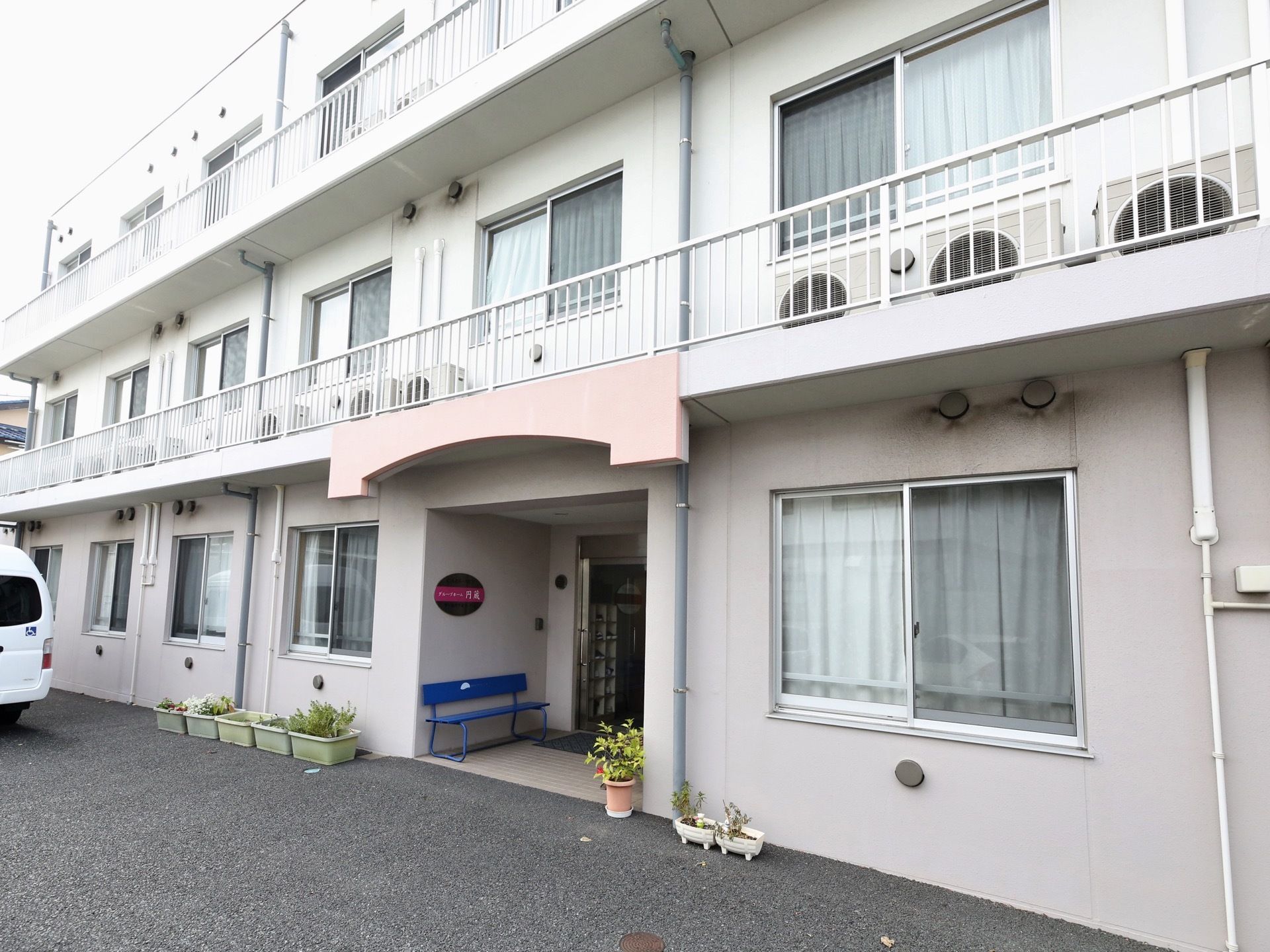 白を基調とした３階建ての集合住宅が建っている。玄関前にはベンチが置かれ、鉢植えの植物が並べられている。